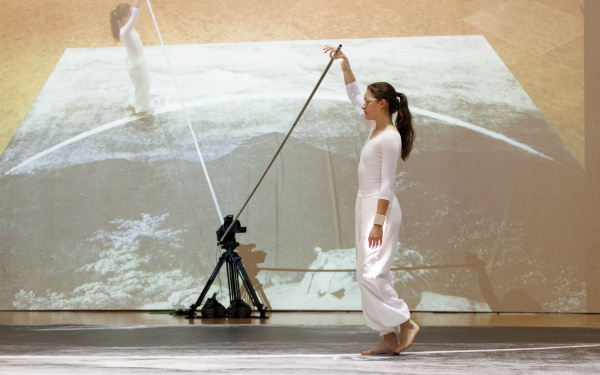 Performance von Ulrike Rosenbachs »Die einsame Spaziergängerin«, dir Künstlerin, ganz in weiß gekleidet hält eine Stange fest und läuft im Halbkreis einen Weg ab, während sie gefilmt wird.