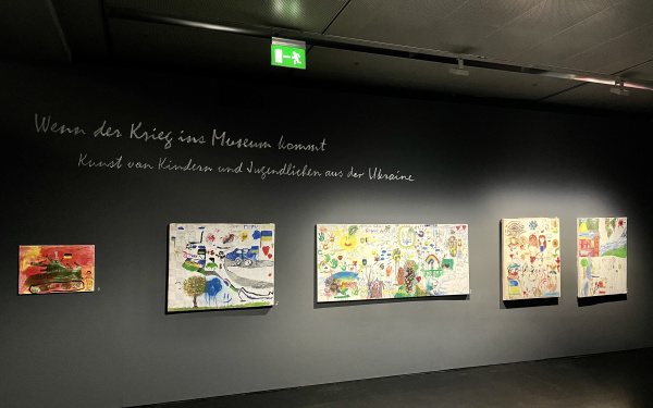Ausstellungsansicht aus der Ausstellung "Wenn der Krieg ins Museum kommt". Zu sehen sind 3 gemalte Bilder an schwarzen Wänden und mit dem Schriftzug: wenn der Krieg ins Museum kommt. Kunst von Kindern und Jugendlichen aus der Ukraine