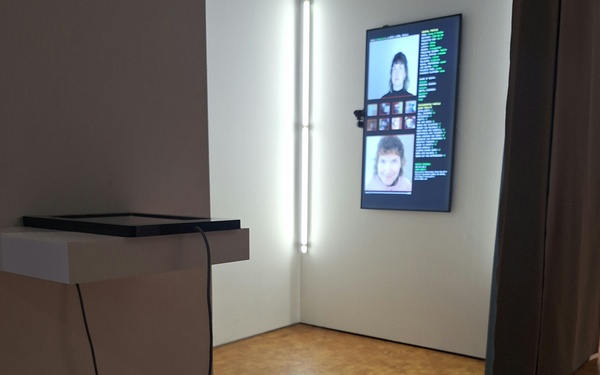 An einer Wand hängt ein Monitor im Porträt. Darauf zu sehen sind ist die wahre Abbildung einer Person und darunter ihre deepfake Version.