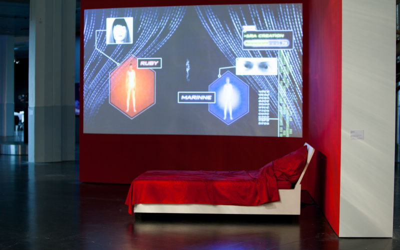 Auf einer Leinwand verschiedene Zeichen. Davor ein Bett mit Samt-Bettwäsche in rot