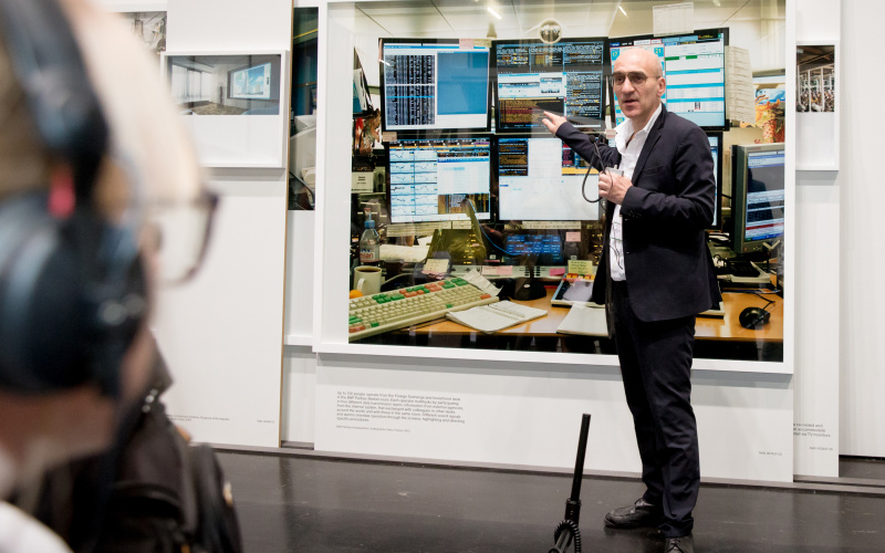 A man stands in front of a picture and talks about it