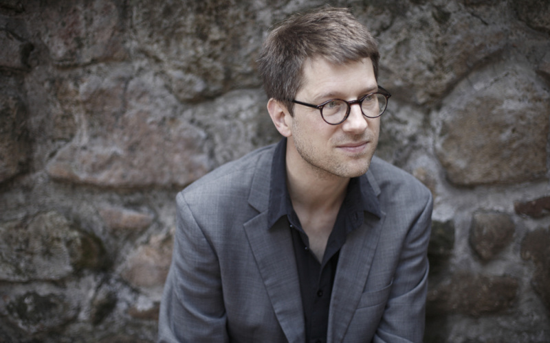 A man with glasses sits in front of a stone wall