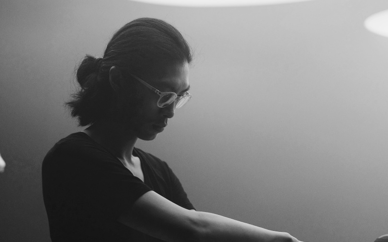 Zu sehen ist ein Mann, welcher im stehen an einem Mixpult steht. Er hat eine Brille und lange Haare, welche er zu einem Zopf zusammen gebunden hat.  