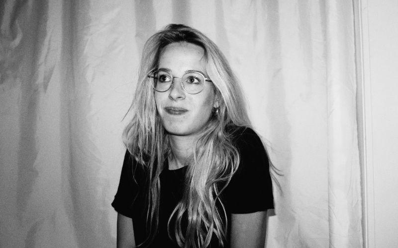 Eine junge Frau mit langen Haaren und einer Brille blickt in die Kamera, sie erscheint fröhlich. Es ist ein Portraitfoto.