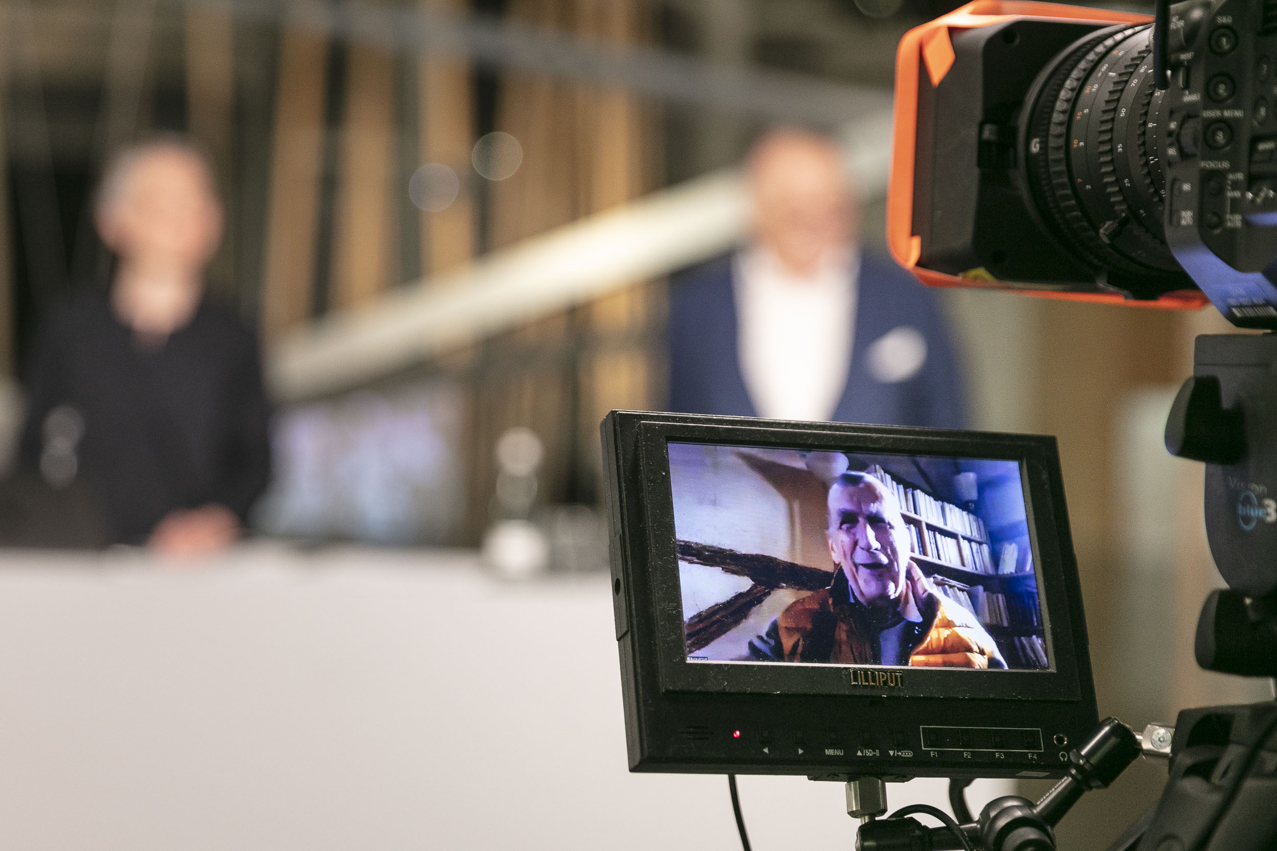 In the foreground is a video camera display with Bruno Latour on it. Behind the display a woman and Peter Weibel can be seen blurred.