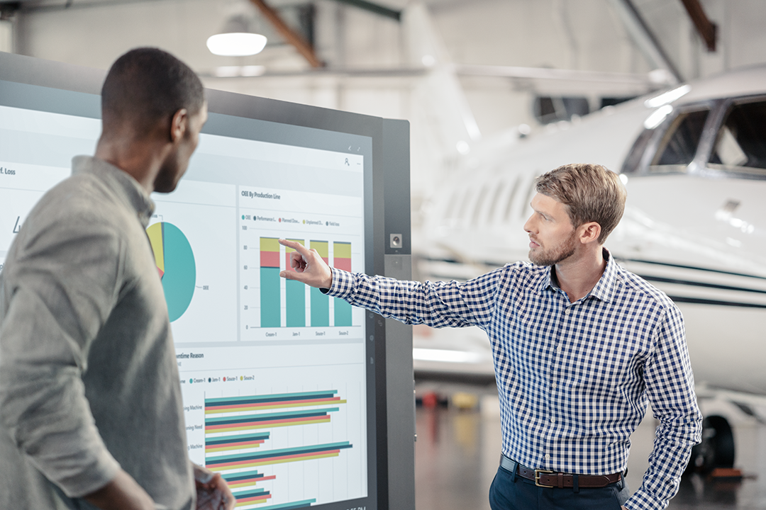 Two men working on a digital screen in a hangar with airplanes