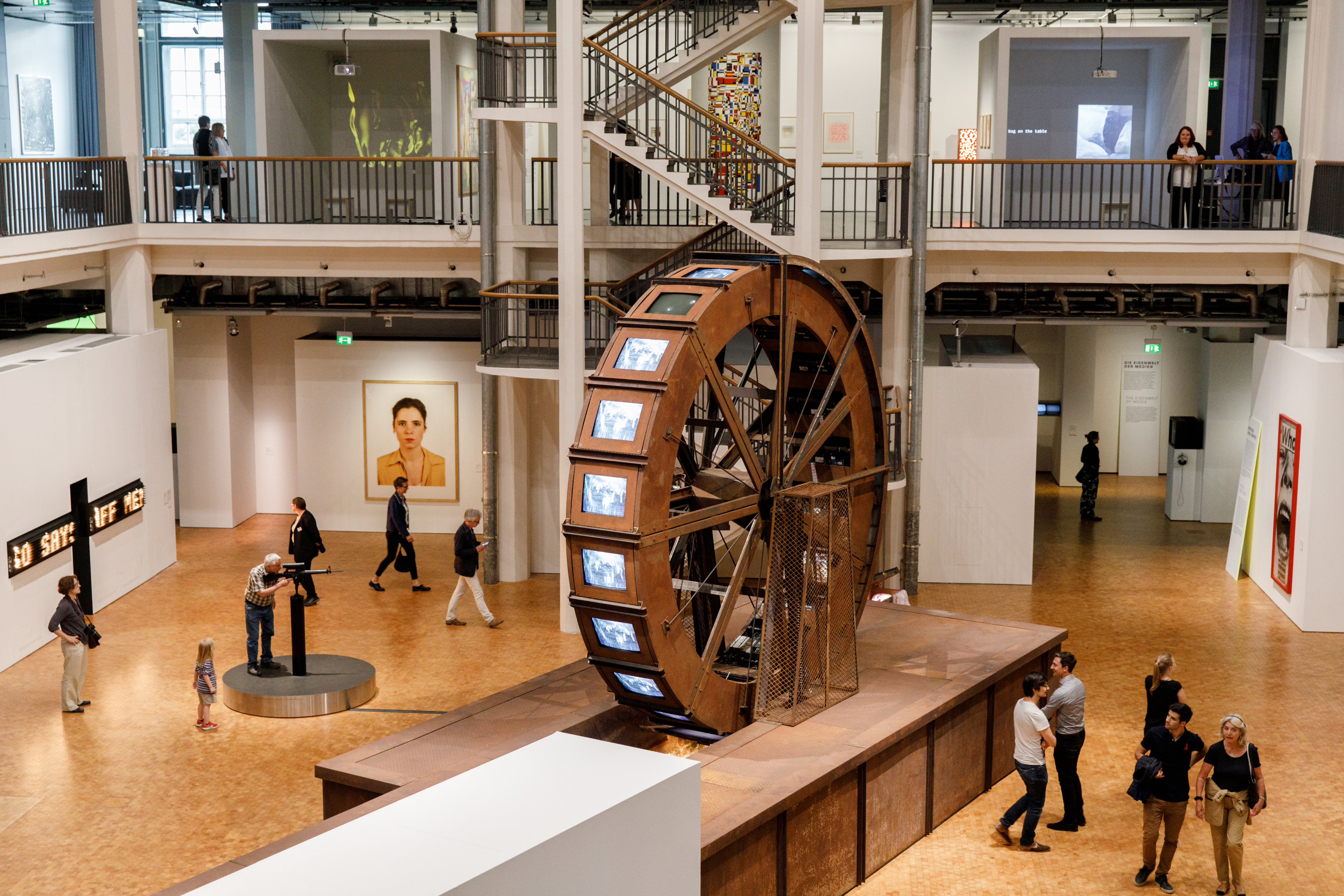 Ein riesiges Mühlrad aus Metall steht in einem Ausstellungsraum. Am Mühlrad befinden sich Röhrenbildschirme worauf Videos von Wasser abgespielt werden.