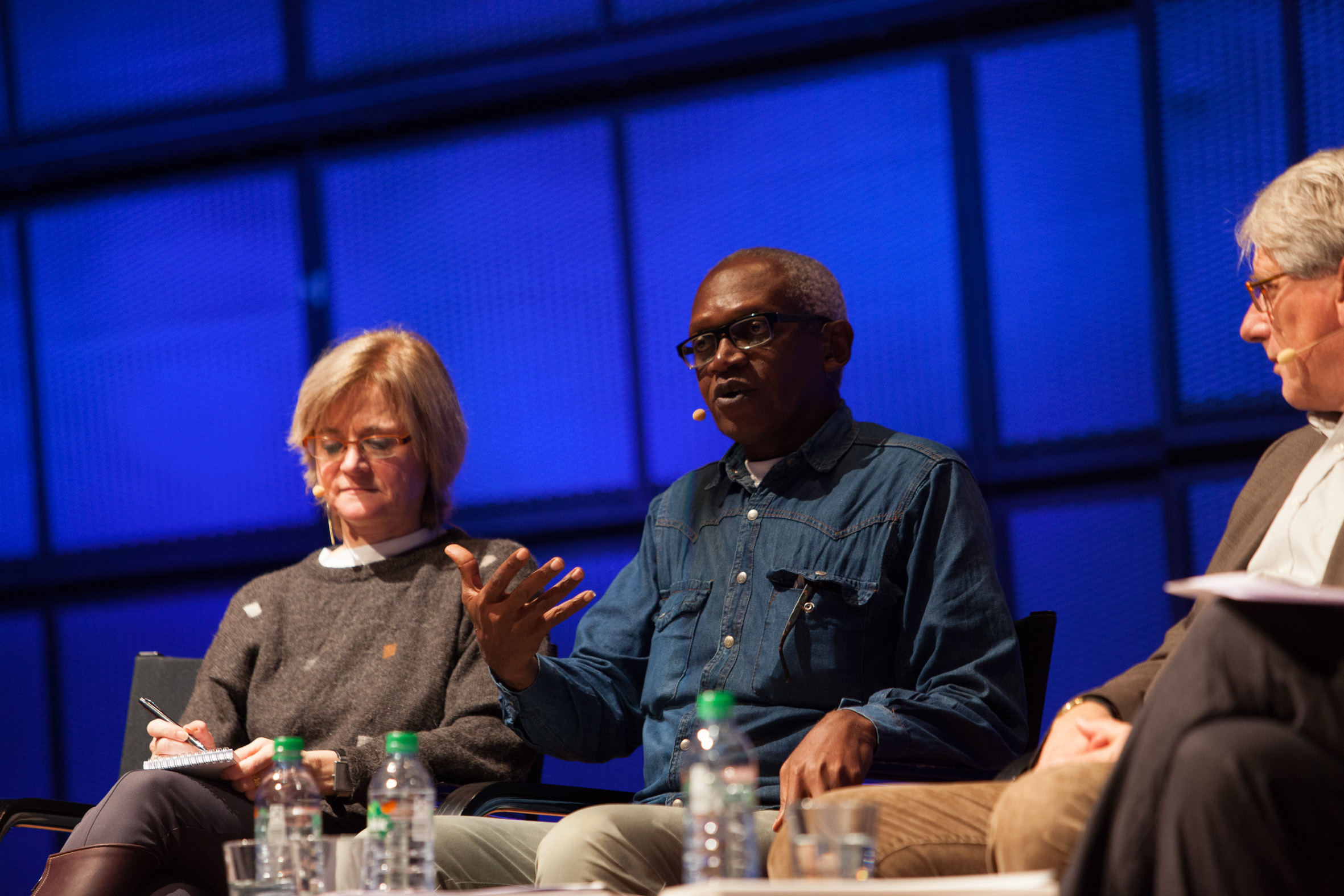 Three people are sitting on a stage while one of them, a man, is talking