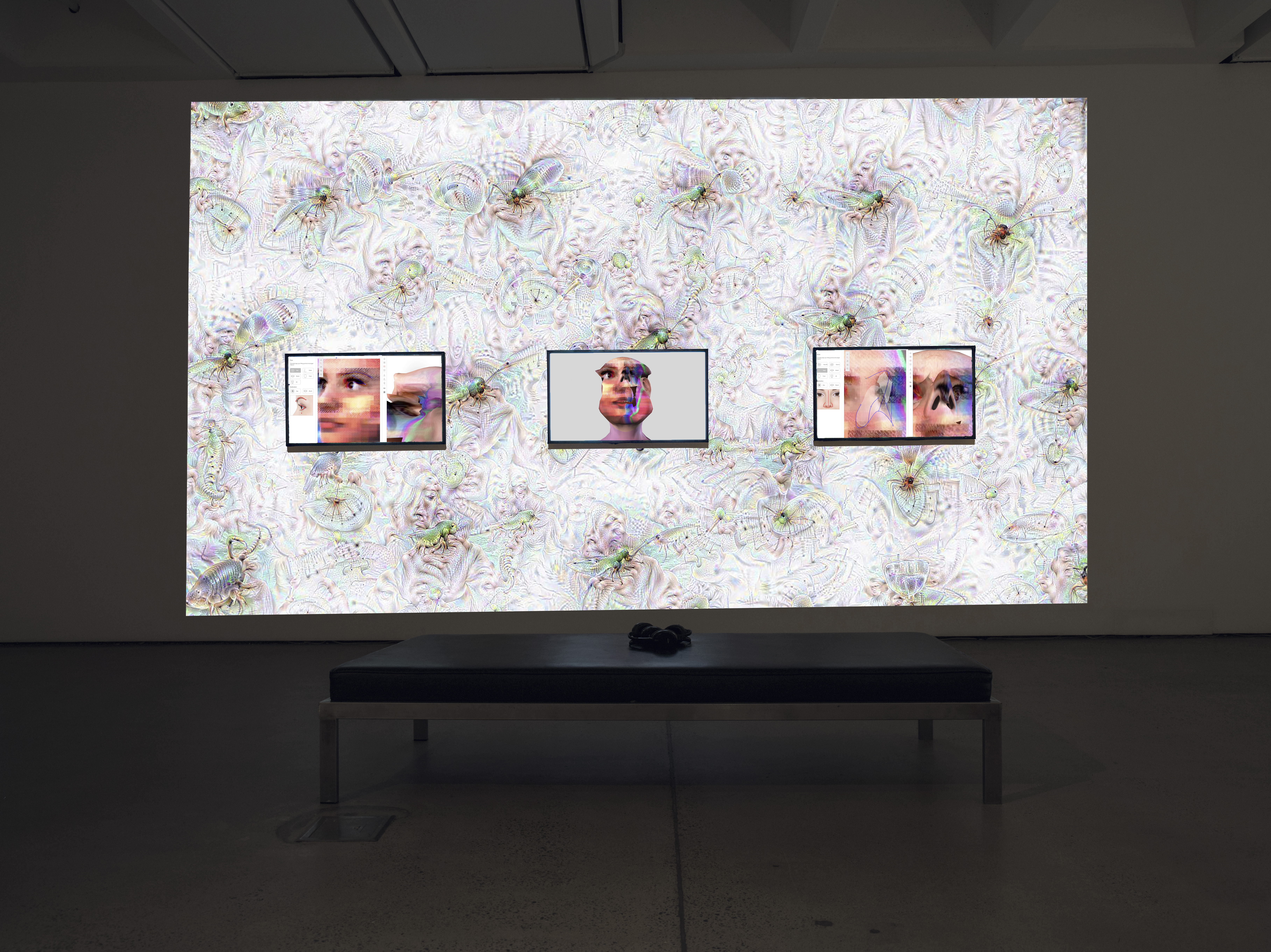 The photo shows an exhibition view of the work »im here to learn so :)))))))«. In the background is a large screen on which a Google DeepDream is projected. Three screens in front of it show the head of the chatbot Tay.