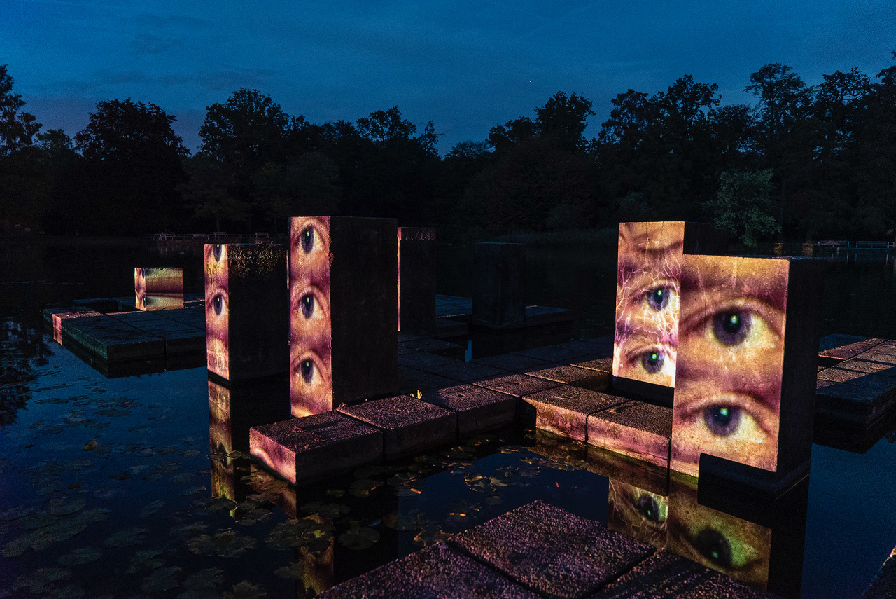 Wasserstelen in einem Park. Es ist später Abend. Auf die Stelen sind Augen gemappt, die offen in die Gegend blicken.