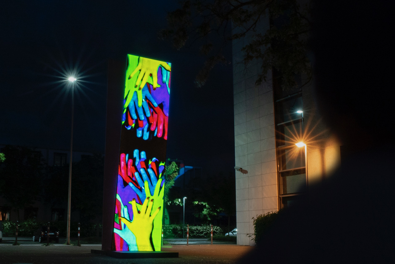 Many hands are projected on a column. One set of hands stretches from bottom to top towards the other set of hands and vice versa. It is night.