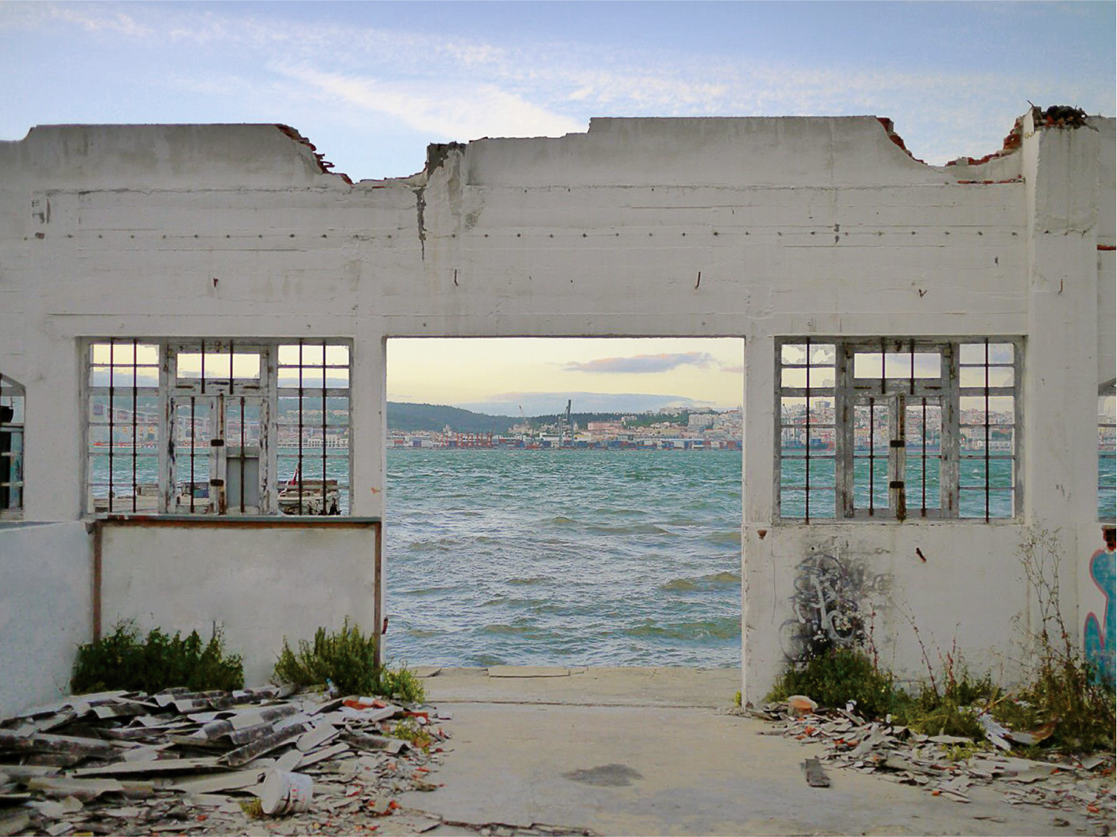 Das Bild zeigt eine Hausruine, durch deren offene Türen und Fenster man auf das Meer und eine dahinter liegende Stadt sehen kann. 