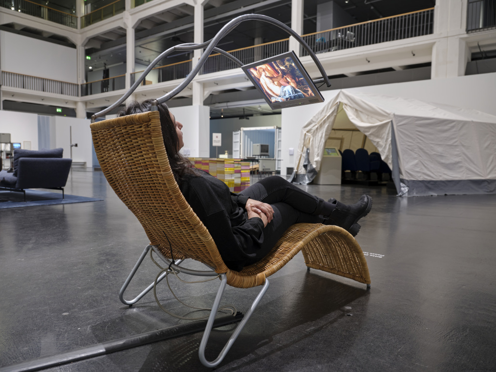 The photo shows a woman lying on a rattan couch. Above the couch is a metal frame which allows a screen to float in the air. On this runs the work Pluriversum by Peter Weibel.