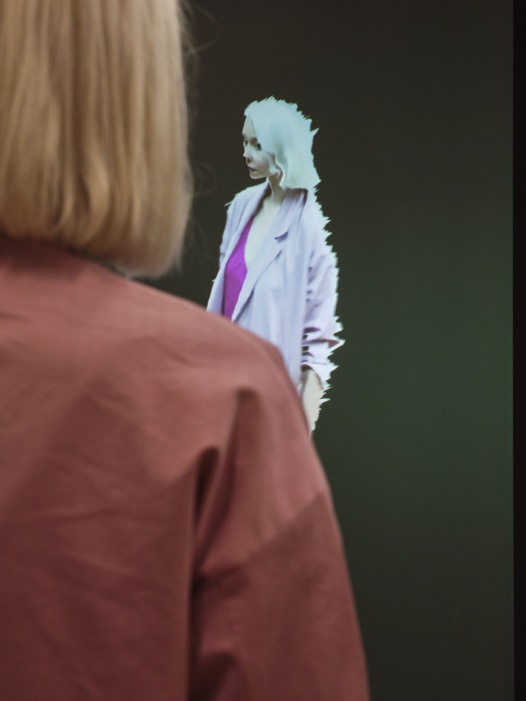 In the foreground of the photo you can see a light blonde woman with short hair and a pink jacket. In front of her she is reproduced as an avatar on a screen. The visitor faces her digital code.