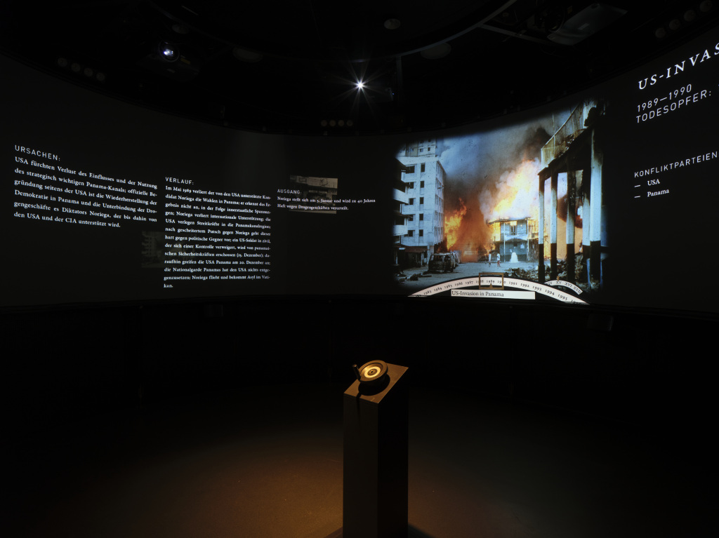 The photo shows a darkened room, which presents war scenarios on a large panorama screen and shows their causes in textform.