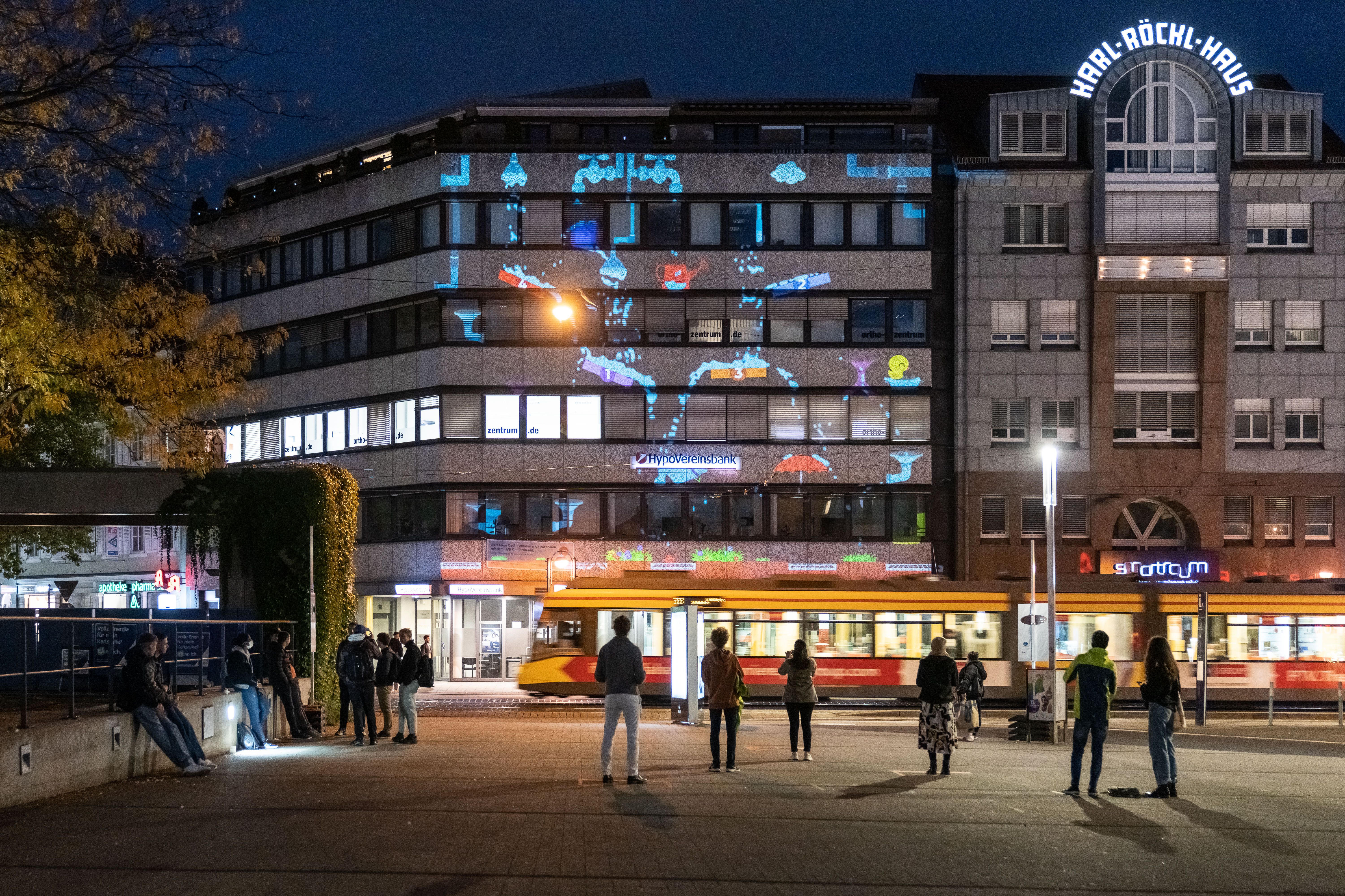 A huge projection on a buildings facade shows individual fab elements distributed: a long faucet, a water gun with funnel, an umbrella.