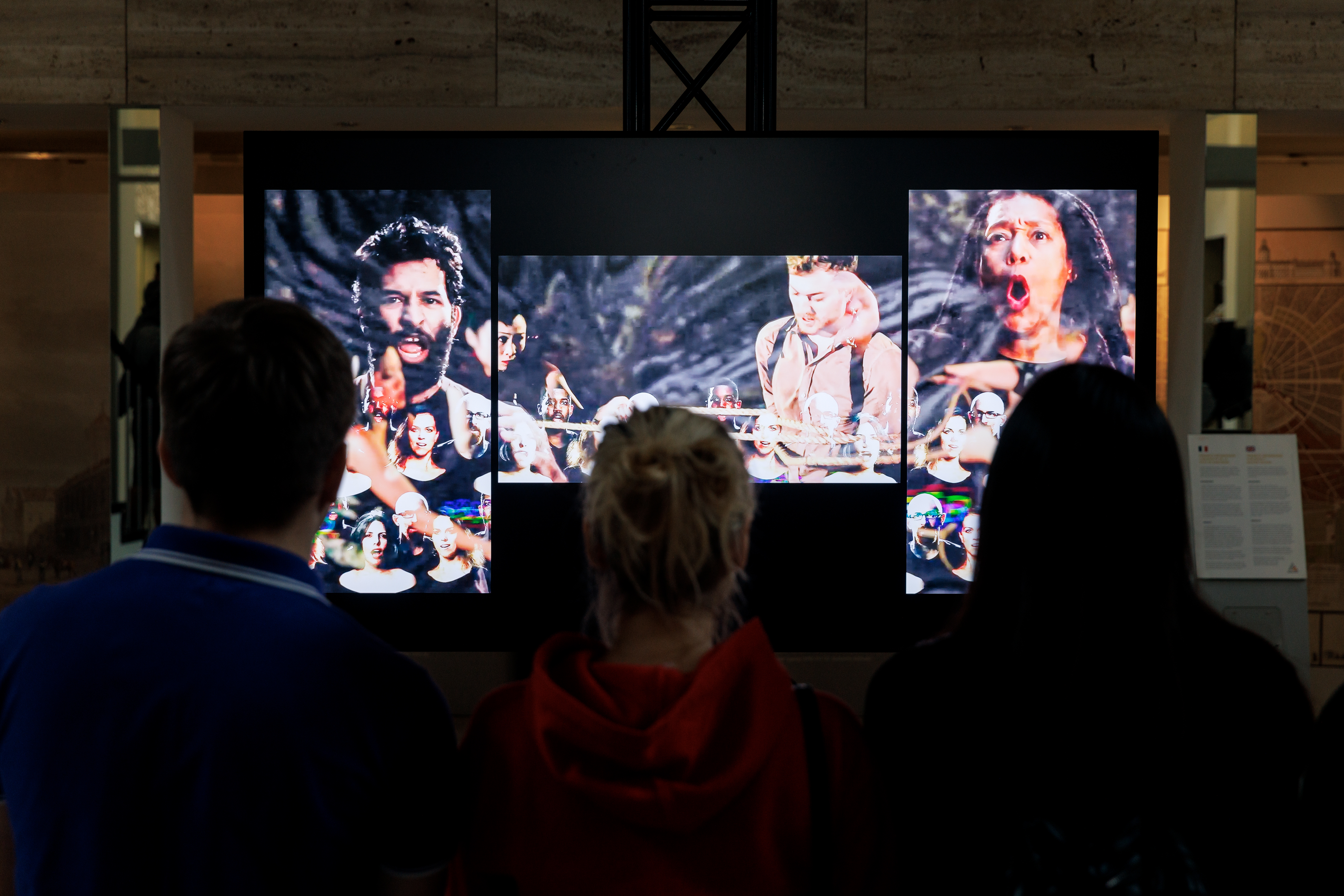 The picture shows the video installation by John Sanborn in the foyer of the rathaus. Aud 3 screens are depicted several people expressing different expressions.