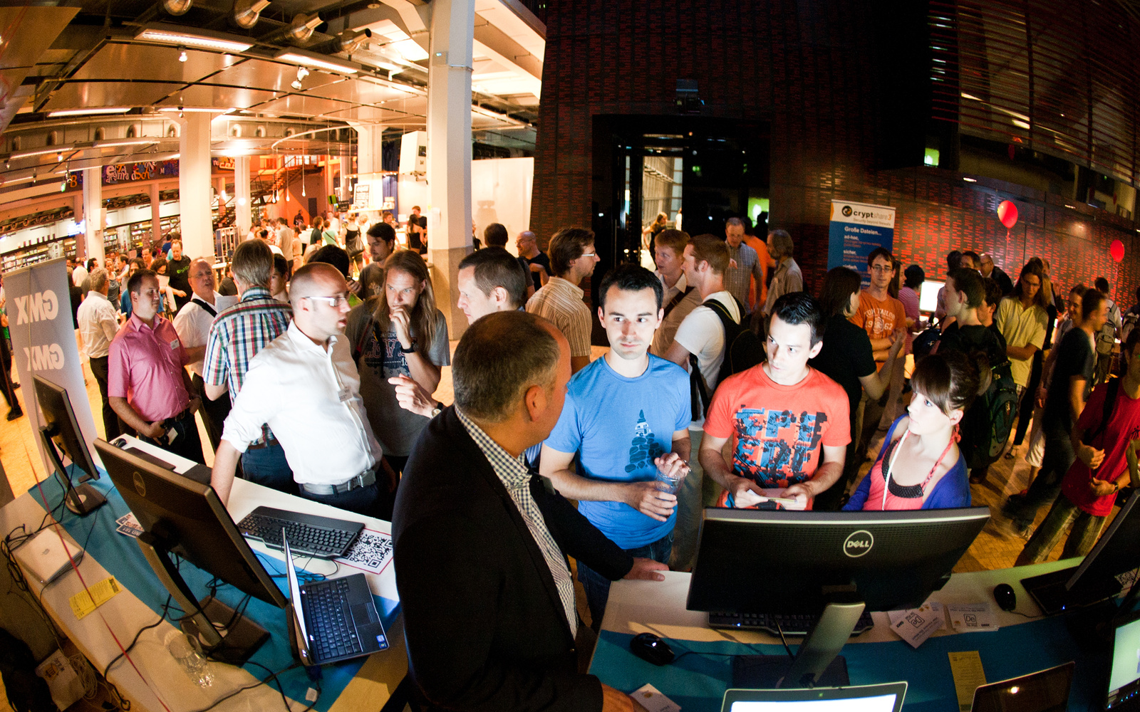 Very many people are in a large hall. In the foreground, computer equipment can be seen.