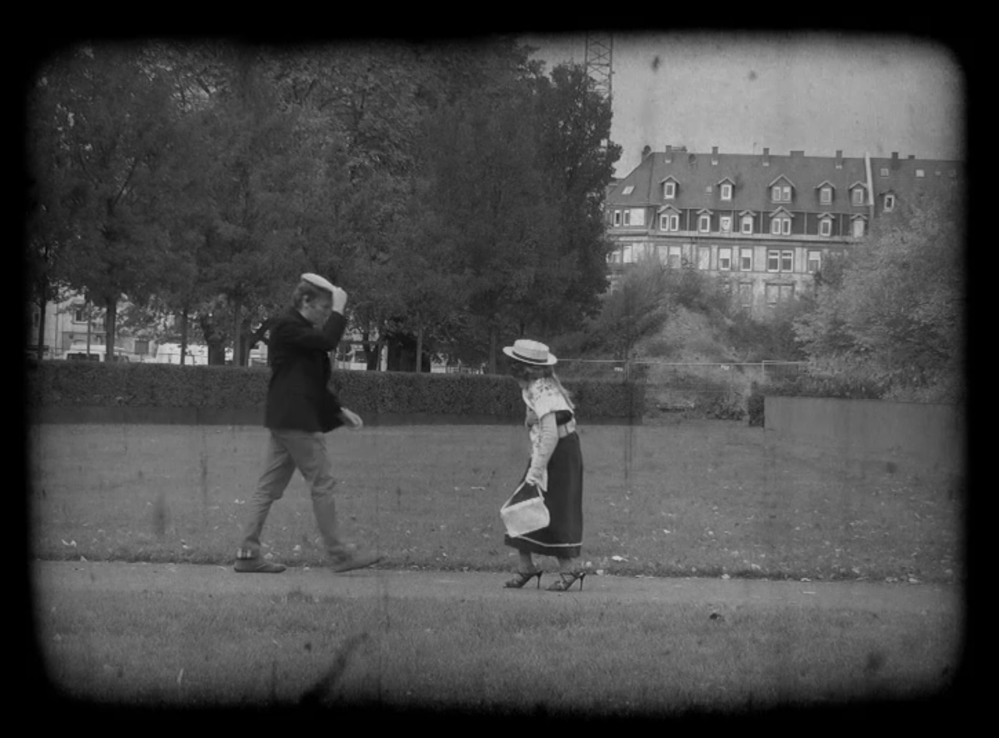 Zwei Passanten begegnen sich auf der Straße, der Herr lüpft den Hut um die sich nähernde Dame zu grüßen. Es ist ein Standbild aus einem schwarzweiß Film, der bei dem Workshop "Die kleinen Strolche" entstanden ist.