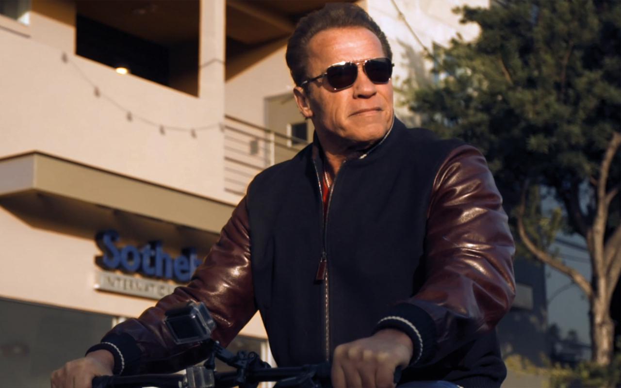 Arnold Schwarzenegger rides a bicycle with sunglasses.
