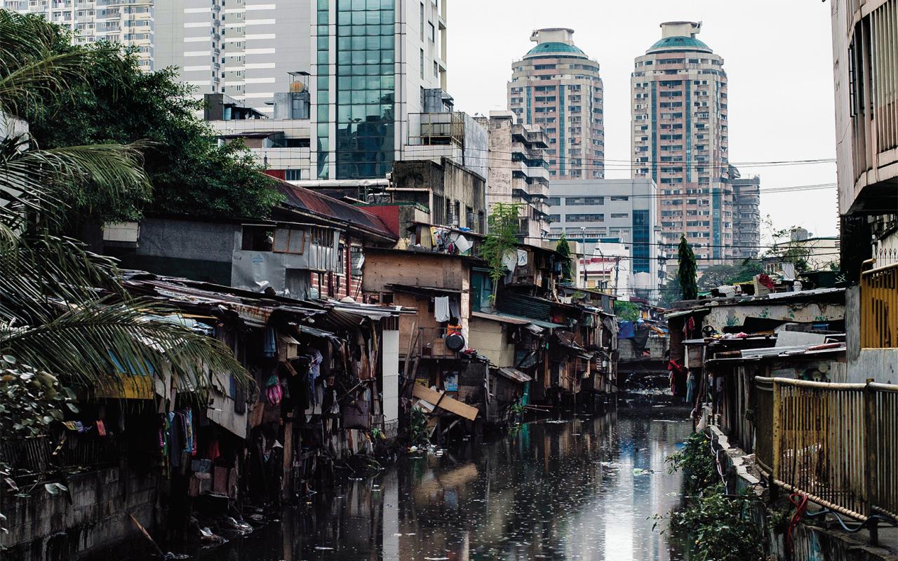 The picture shows dilapidated huts on a dirty river in front of modern high-rise facades. 
