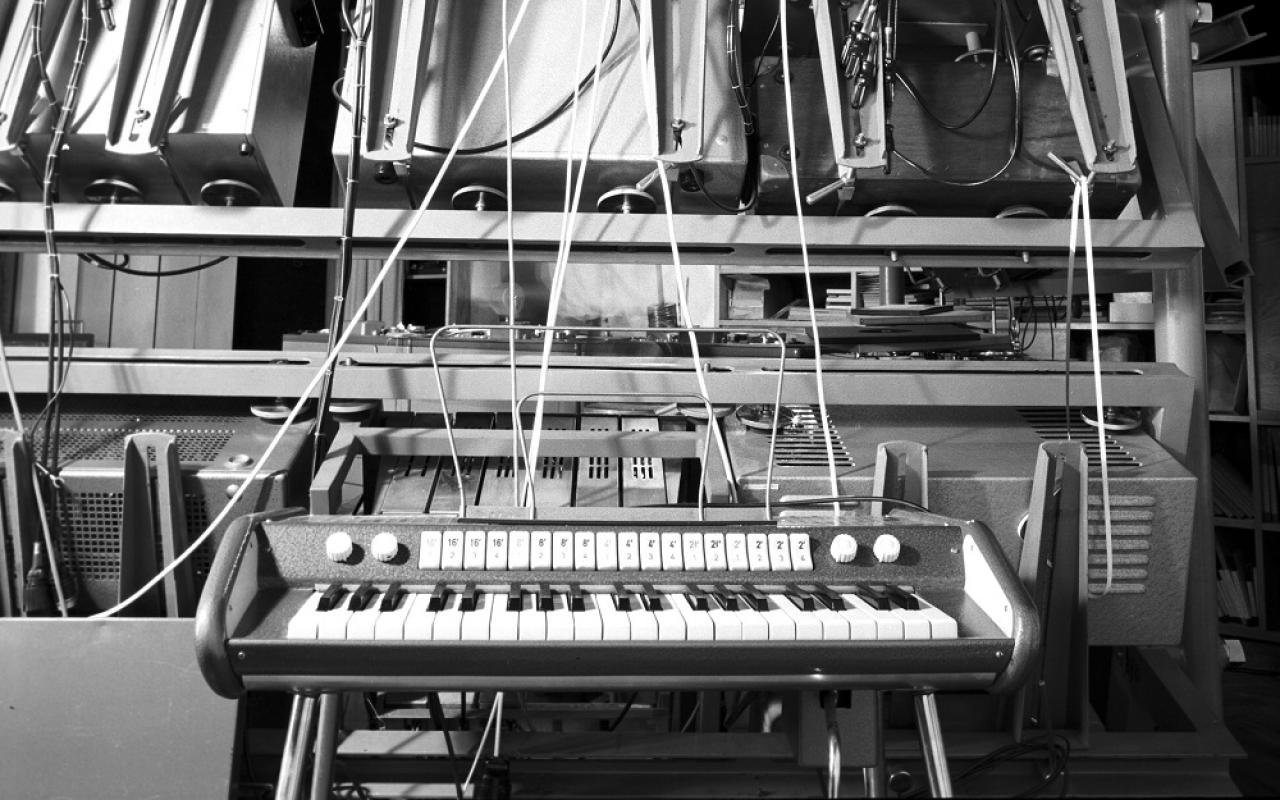 A black and white picture shows an electric piano in front of cables and computers. 