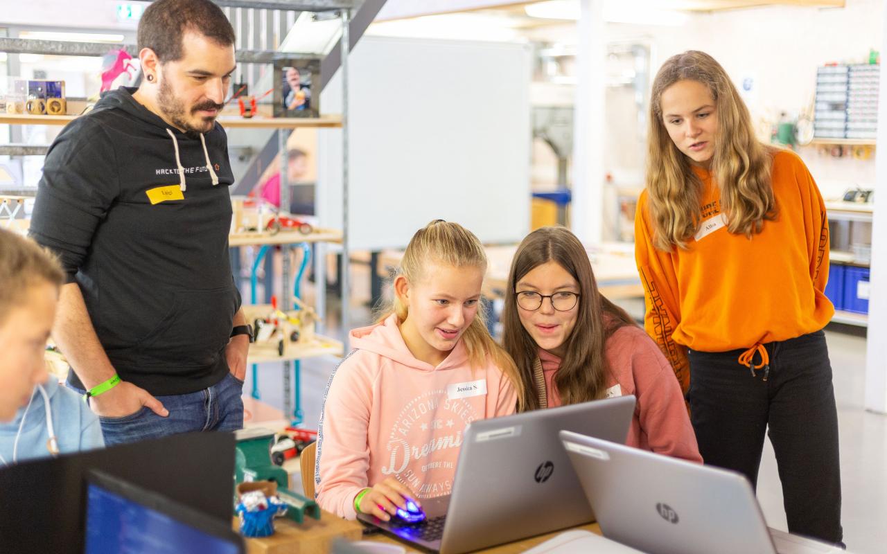Ein Foto der Veranstaltung Hack to the Future, zwei junge Teilnehmerinnen schauen auf ihre Laptops, daneben stehen eine Mentorin und ein Mentor.