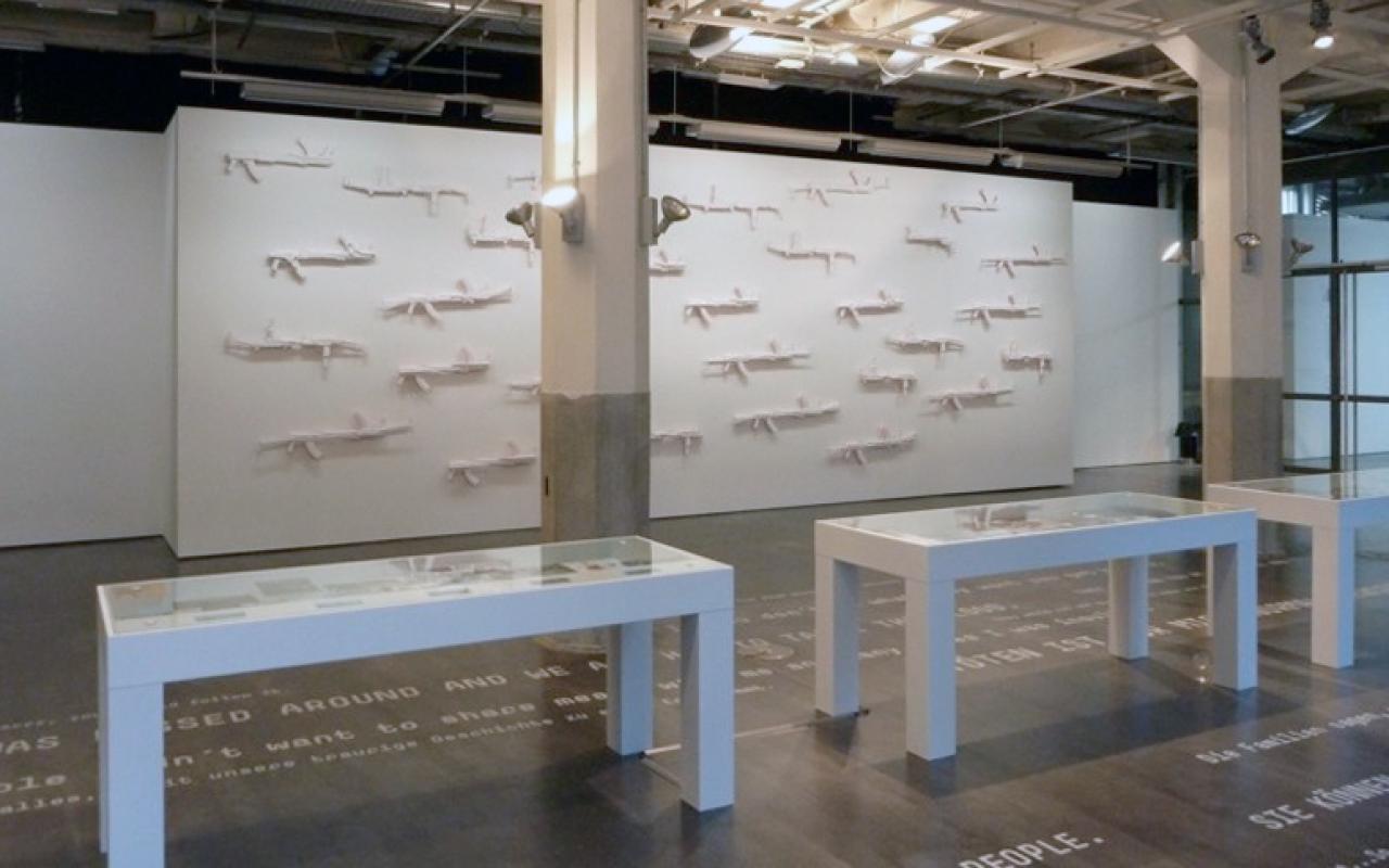 View of the exhibition »Symbiosis«: In the foreground, white cabinets, in the background on the wall: white guns