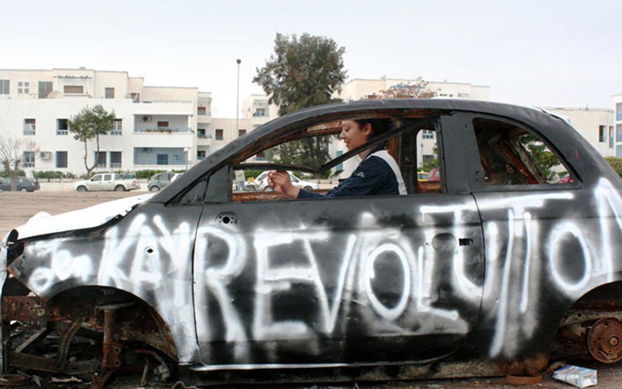 A woman sitting in a non-functioning car. On the driver's side was sprayed with white color "revolution".