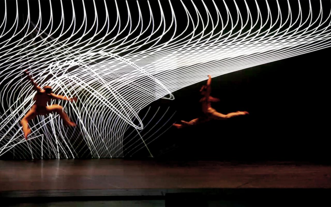 Eine Bühne. Zwei Tänzer bewegen sich durch die Lüfte. Im Hintergrund spiralartig weiße Gebilde.