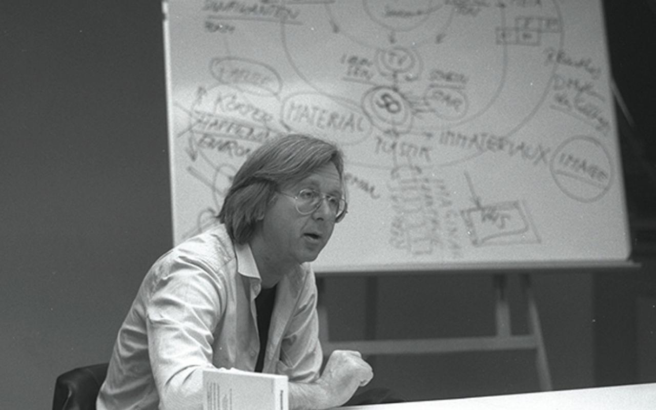 Black and white photograph of a man sitting at a table. In the background a flip chart.
