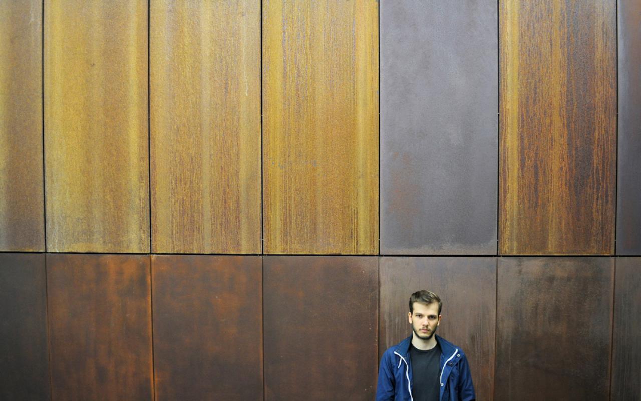 A young man wearing blue sports jacket standing in front of a wooden wall