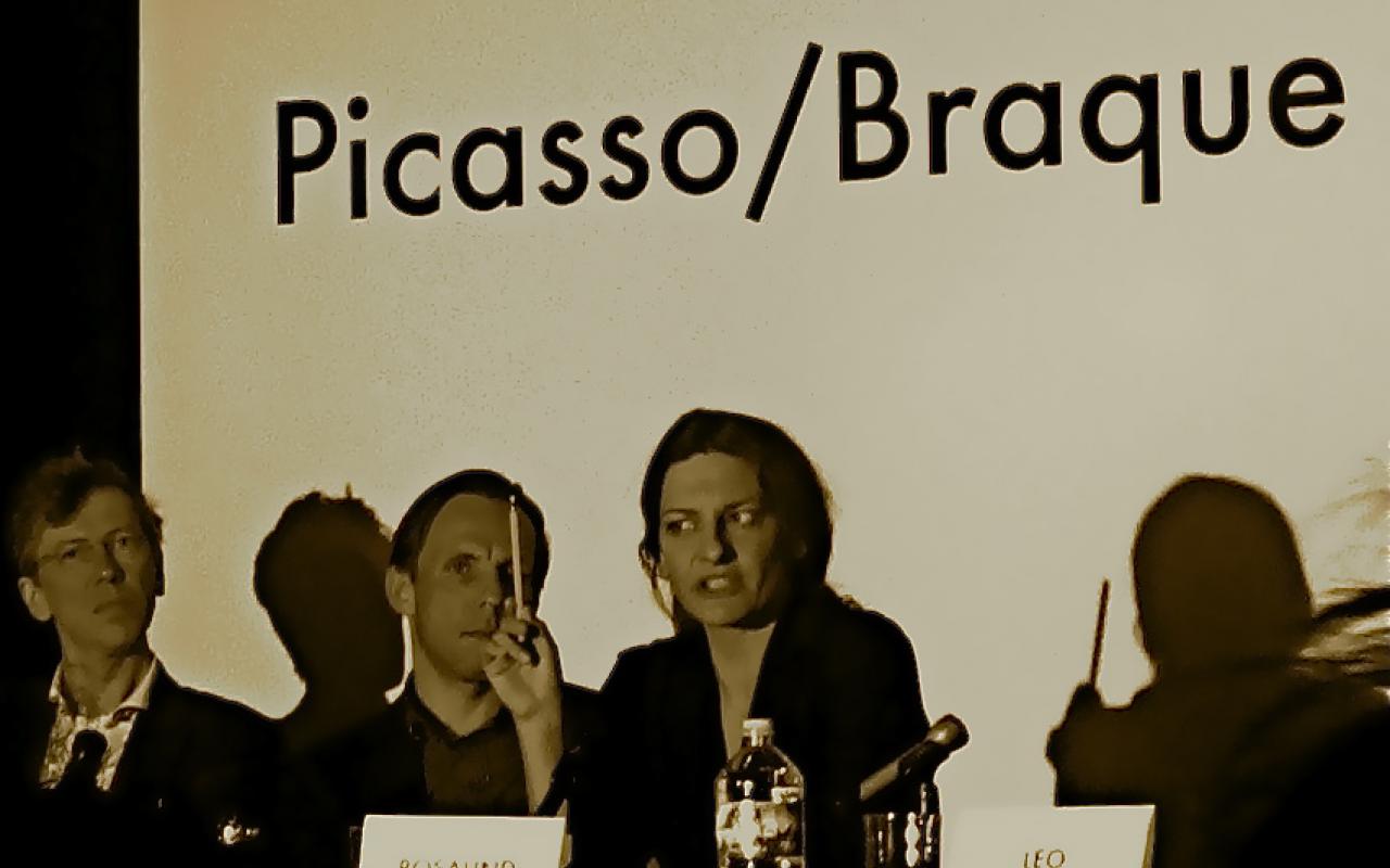 From left to right: Two men and a woman at a table. She talks, keeping a pencil in her right hand. On the table name tags. In the background is 'Picasso / Braque' to read.