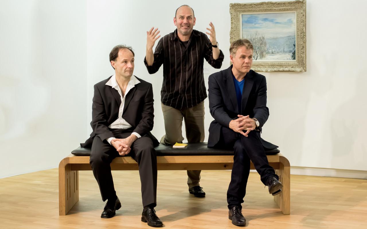 Three Men in a museum. Two sit and look disinterested to the left. One of them kneels. He raises his arms and looking with a desperate expression directly into the camera. Behind the men is a painting. It shows a snowy landscape.
