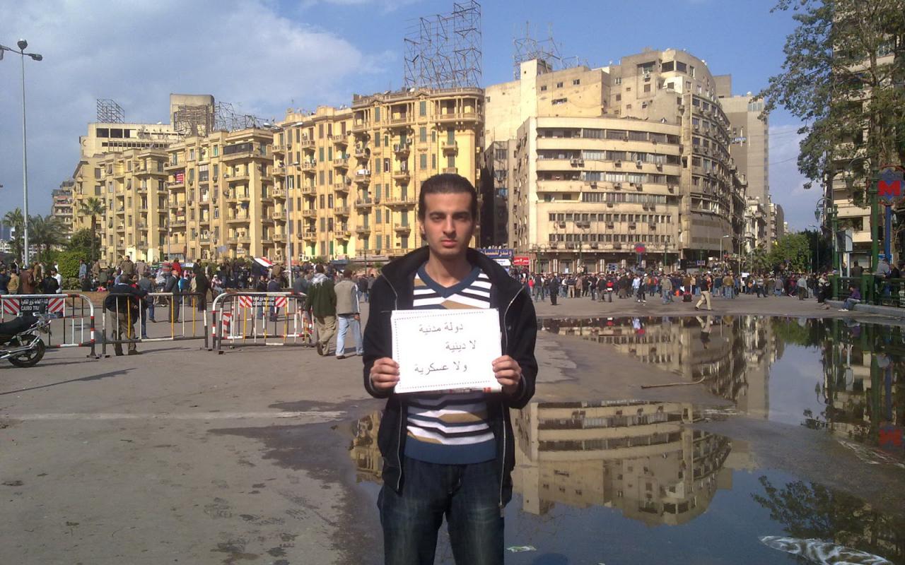 Ein junger Mann hält eine Botschaft in ägyptischer Sprache in seinen Händen. Im Hintergrund ist ein architektonisches Stadtbild zu erkennen.