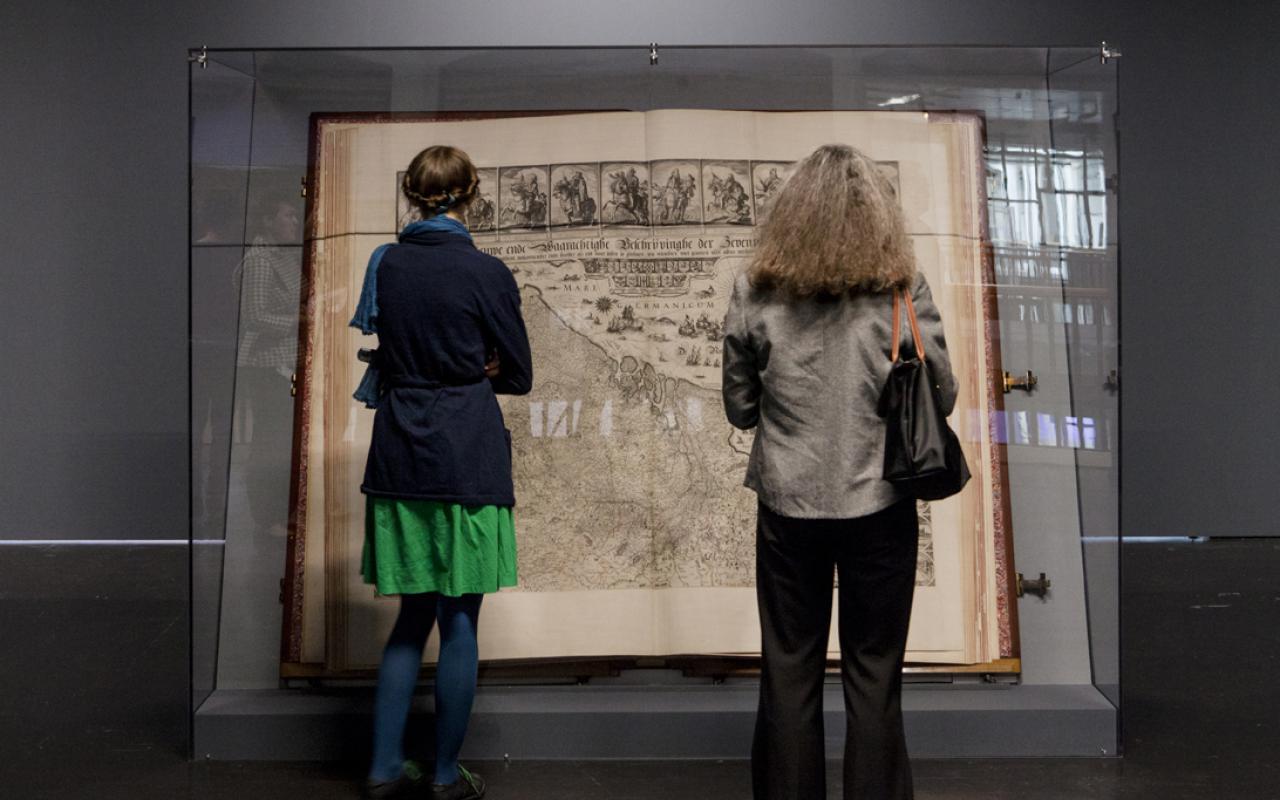 The Klencke Atlas (1660) is one of the largest atlas in the world. Aerated he stands in a glass structure that protects it. Two women seen from behind looking at it.
