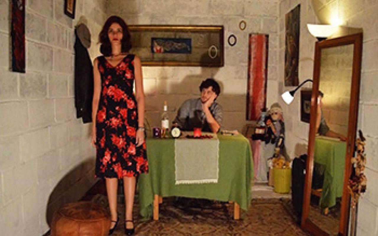 View in room with mirror and table. This has a green tablecloth. On the table sits a man. Next to him stands a woman in a black red floral dress.