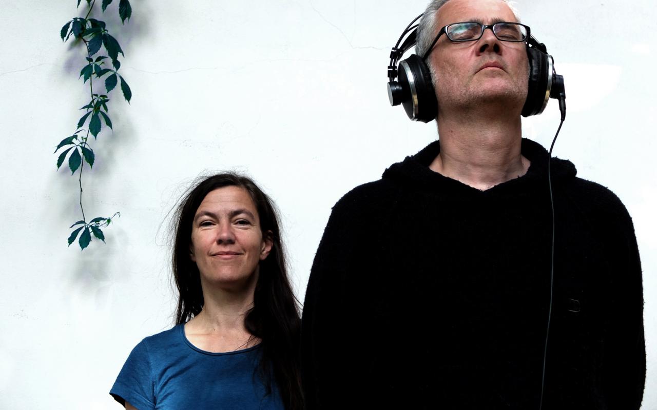 A man with closed eyes and headphones stands right in front of a woman who looks at the camera