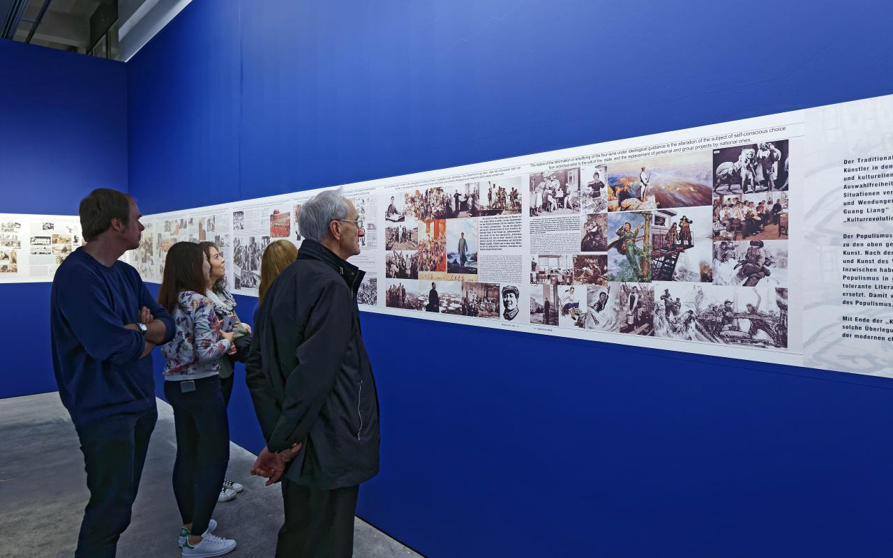 Fünf Menschen betrachten Bilder in einer Ausstellung, die auf blauem Grund hängen