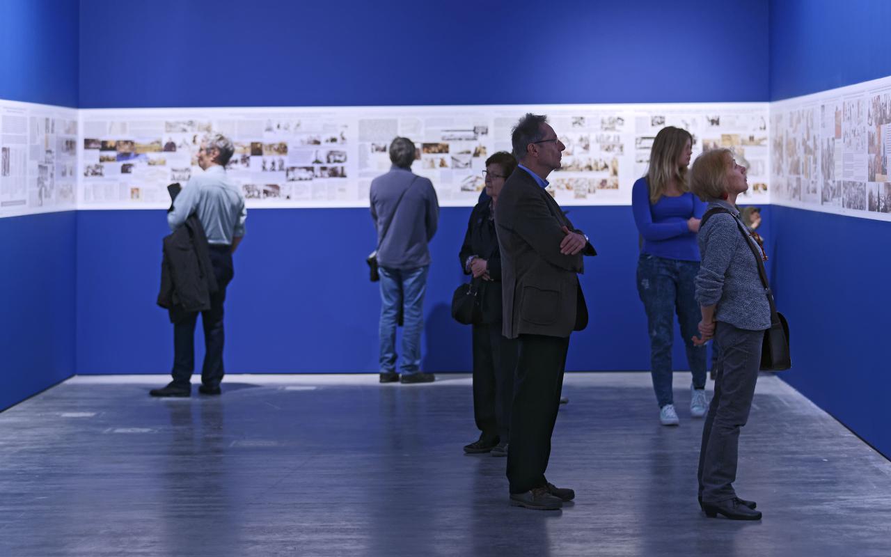 Menschen sehen sich Bilder der Ausstellung an, die auf blauem Hintergrund ausgestellt sind