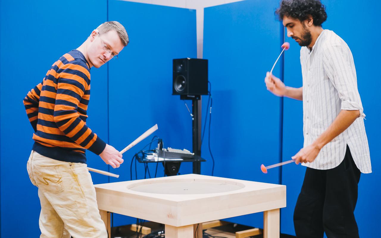 Two men with mallets stand at a table which has a round plate in the middle.
