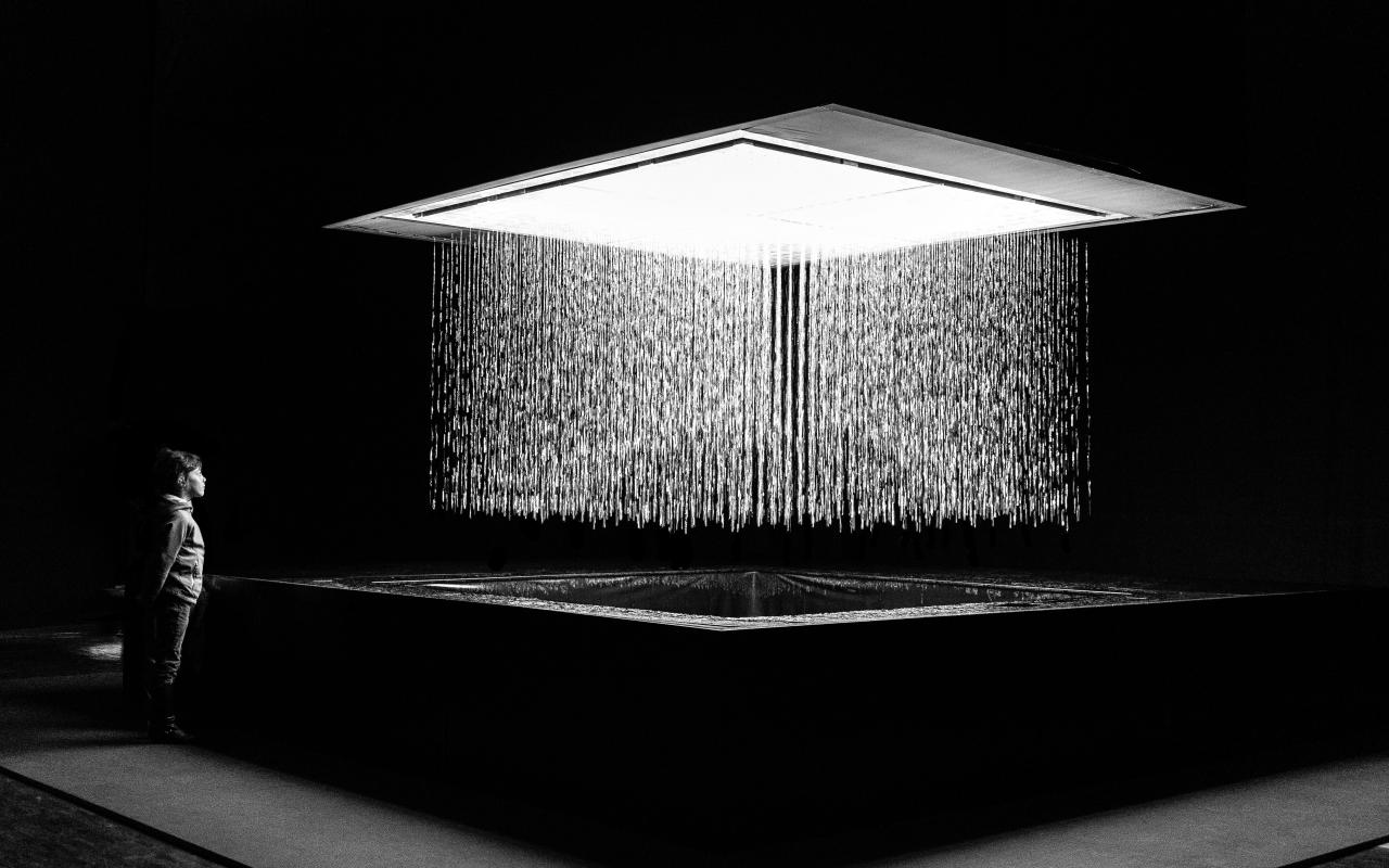 Black and white photo of the installation 3D WATERMATRIX in which water falls to the ground and generates 3D shapes. A person is watching the insatallation