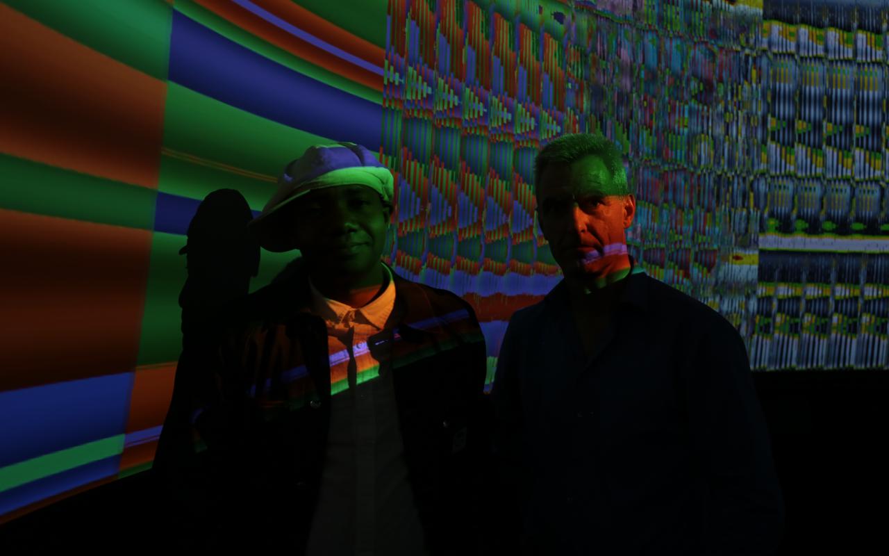 Zwei Männer stehen in einem dunklen Raum vor einem Screen mit bunten Farben.