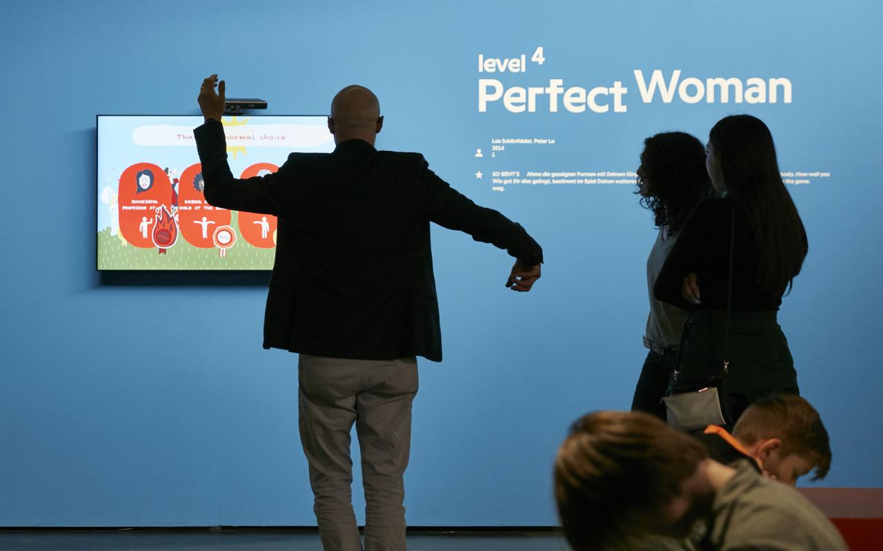 Vor einer blauen Wand, an der ein Bildschirm befestigt ist, steht ein Besucher mit erhobenen Armen. Weitere Zuschauen stehen betrachtend daneben. 