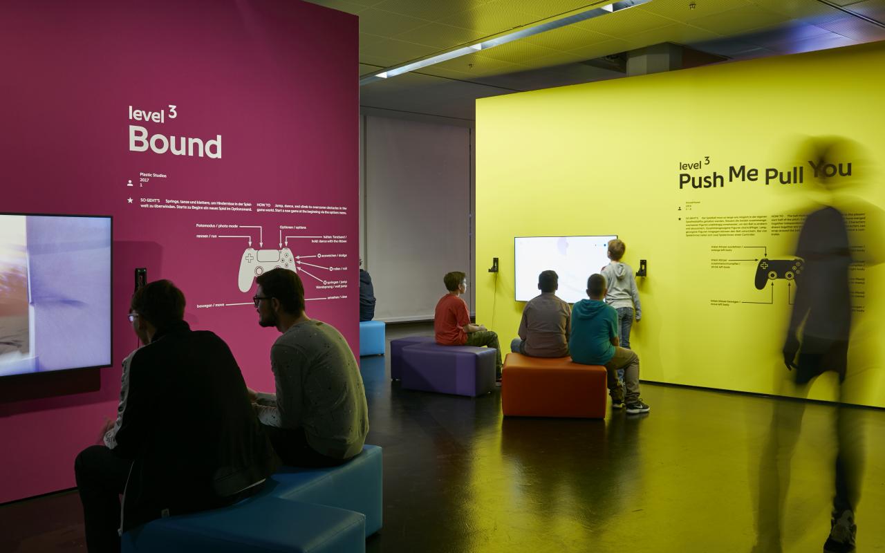 Links im Vordergrund sitzen auf einer Sitzbank zwei Besucher vor einem Bildschirm, der an einer magentafarbenen Wand befestigt ist. Rechts etwas im Hintergrund sitzen mehrere junge Besucher und Besucherinnen vor einer gelben Wand mit Bildschirm. 