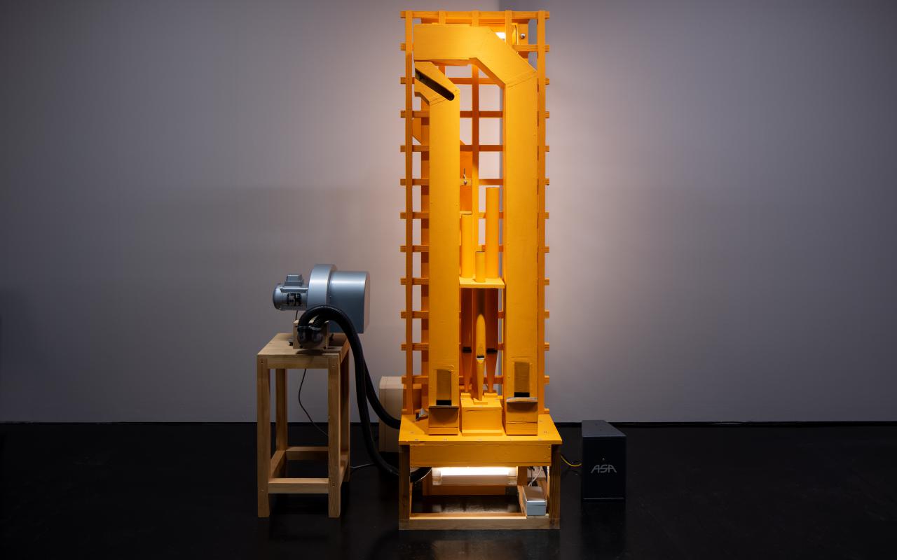 On display is a rectangular, machine-like installation standing on a stool. To the left of the installation there is also a small stool on which an object lies. 