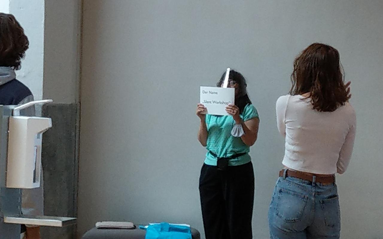 Carmen Beckenbach hält ein Schild vor ihr Gesicht auf dem der Workshop Silent Action angekündigt wird.