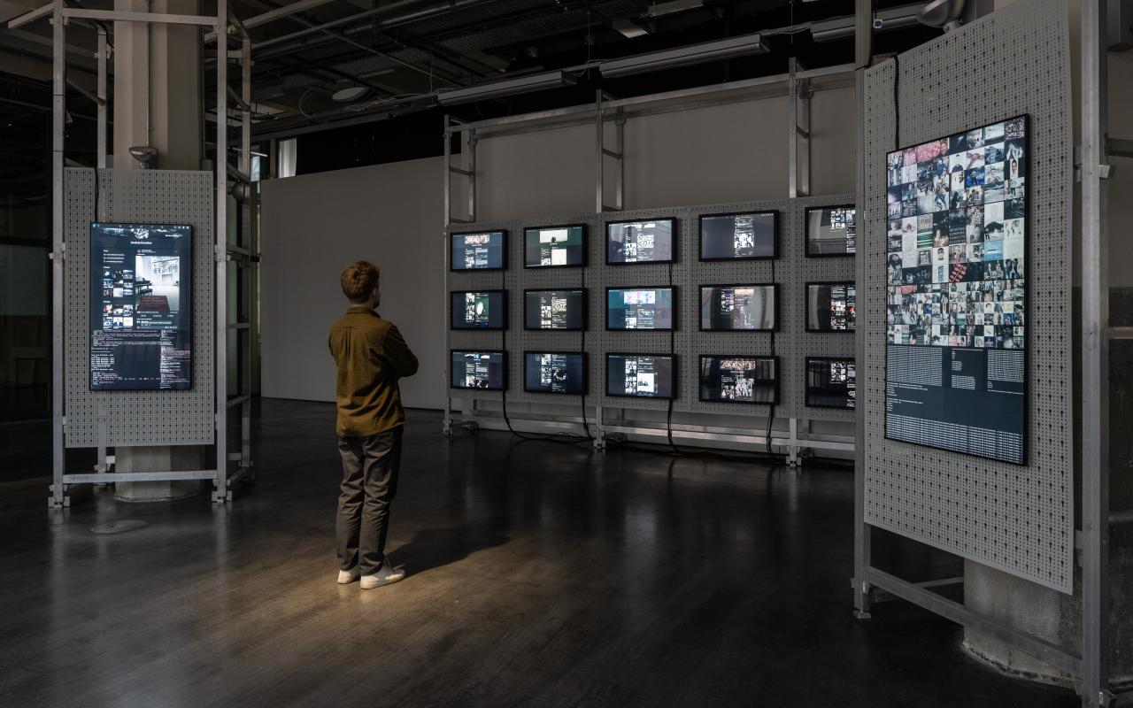 You can see a large open space with a man standing in the middle looking at many screens. The screens hang on the wall and display many different codes and shapes. 