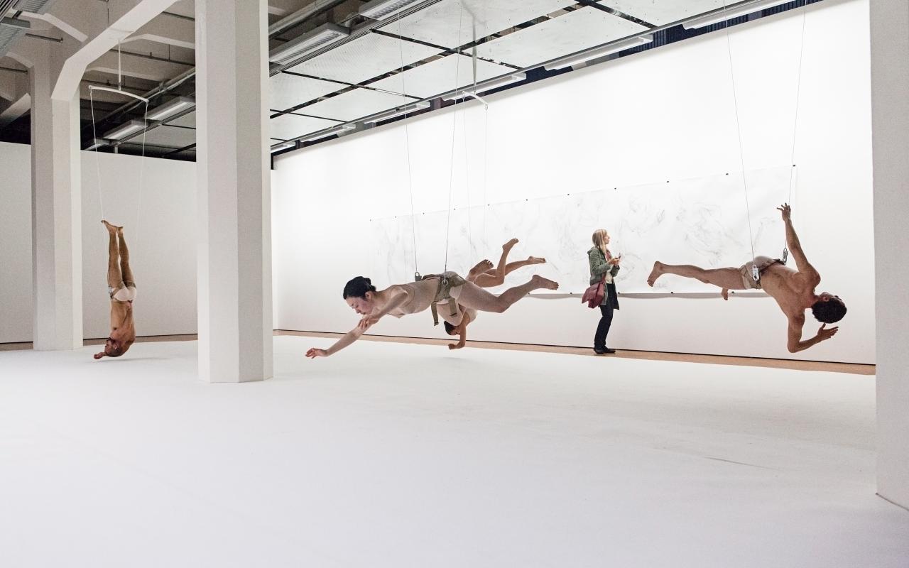 Fünf Tänzer hängen an Drahtseilen kopfüber im Ausstellungsraum.