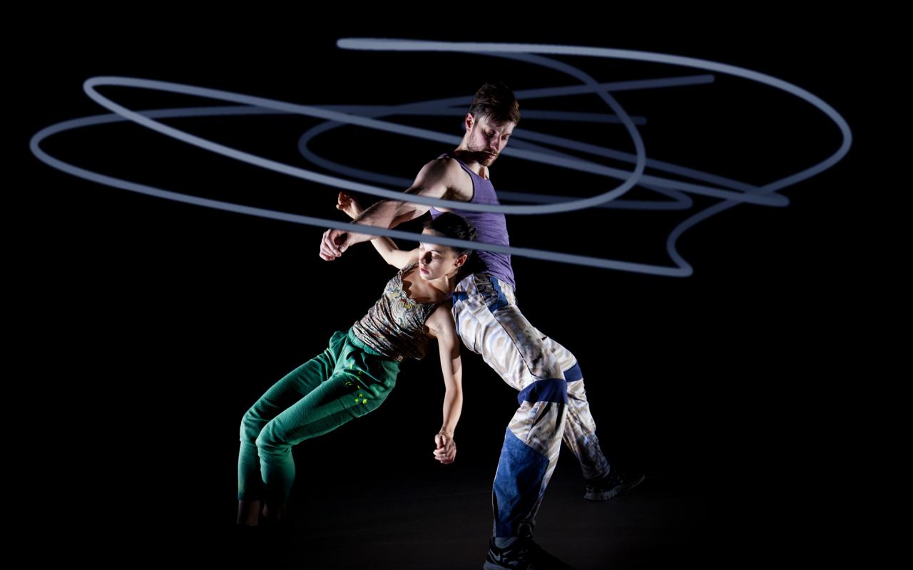 Zwei Tänzer vor schwarzem Hintergrund, umhüllt von kreisrunden Lichtkunststrahlen